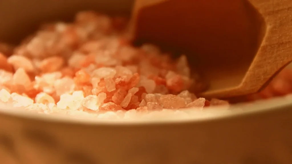 A jar of coarse sea salt body scrub with green tea leaves on a wooden spa tray.