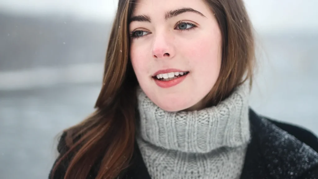 A woman in a knit hat and scarf in the snow applying protective cream to her cheeks.