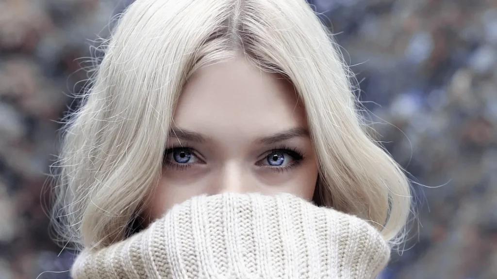 A woman in a cozy oversized wool sweater, with a stack of knitted sweaters and turtlenecks beside her.