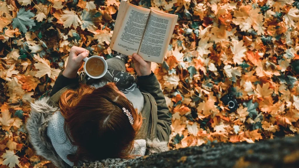 A smiling woman reading a book under a blanket with tea on an autumn evening.