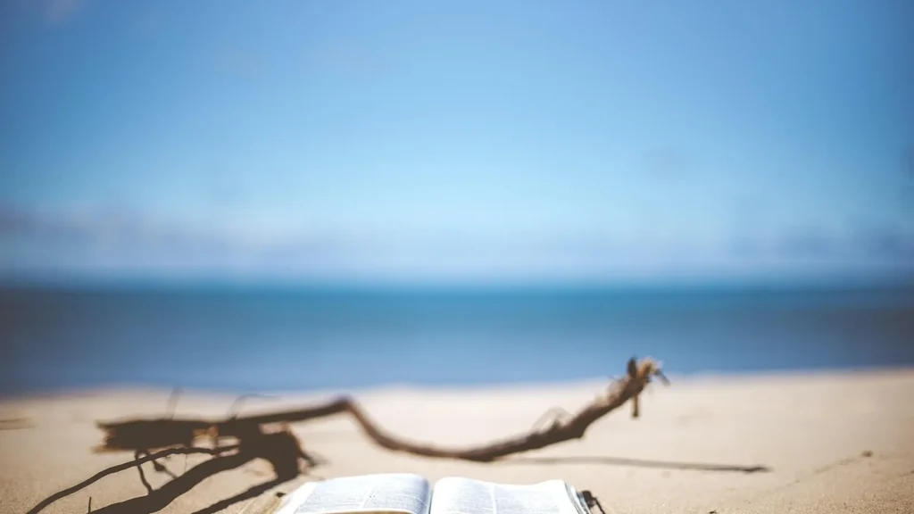 A woman reading a book on a sunny beach lounger with sunglasses and a hat.